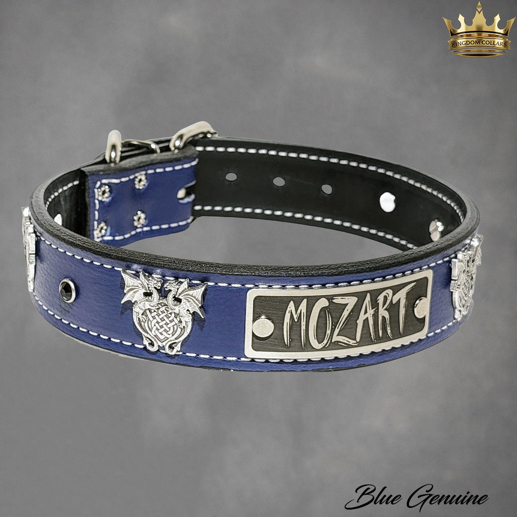 A blue leather dog collar with silver metal hardware, featuring a central rectangular plate engraved with 'MOZART', a winged emblem on the left, and white contrast stitching along the edges. The collar is displayed on a textured gray background with a gold brand logo in the top right corner.