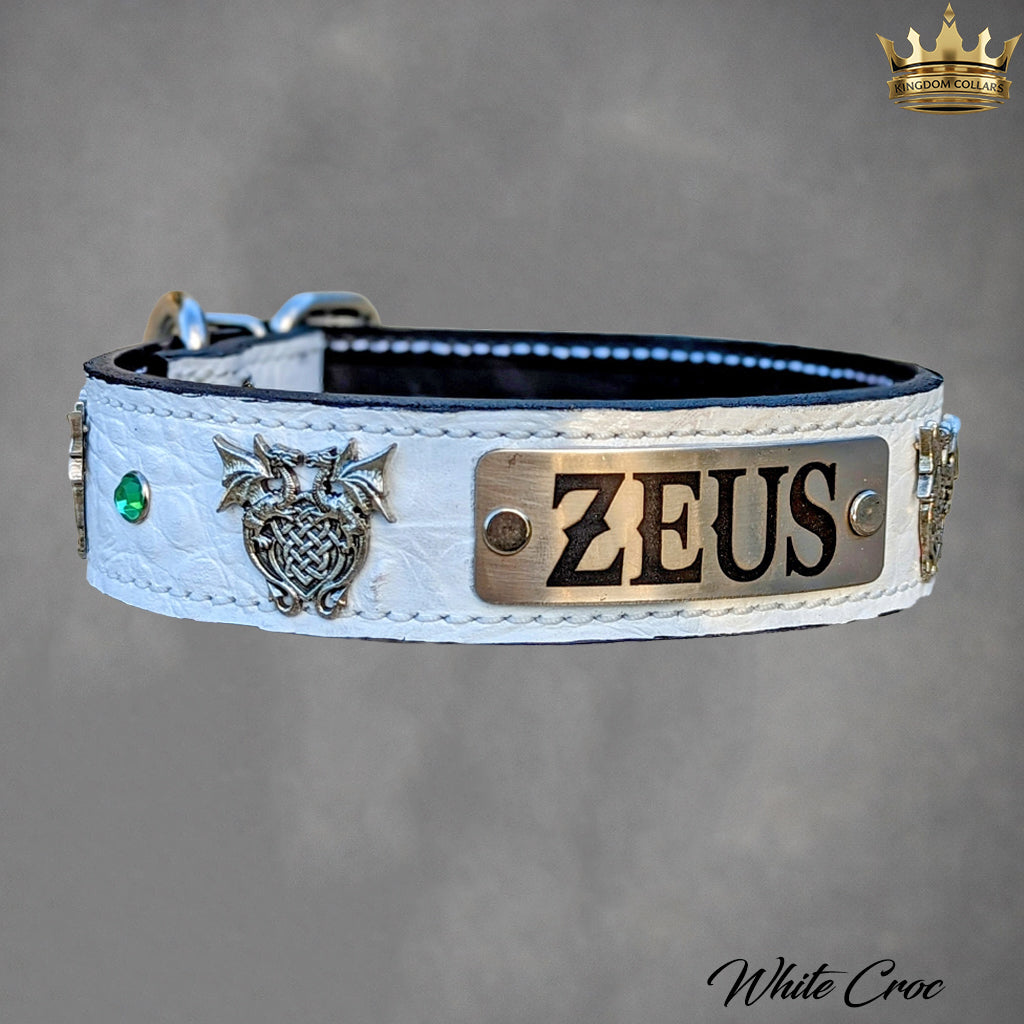 A white crocodile-embossed leather dog collar with a silver-toned metal plaque engraved with 'ZEUS', a silver owl charm, a small green stone, and silver hardware, shown on a gray background.