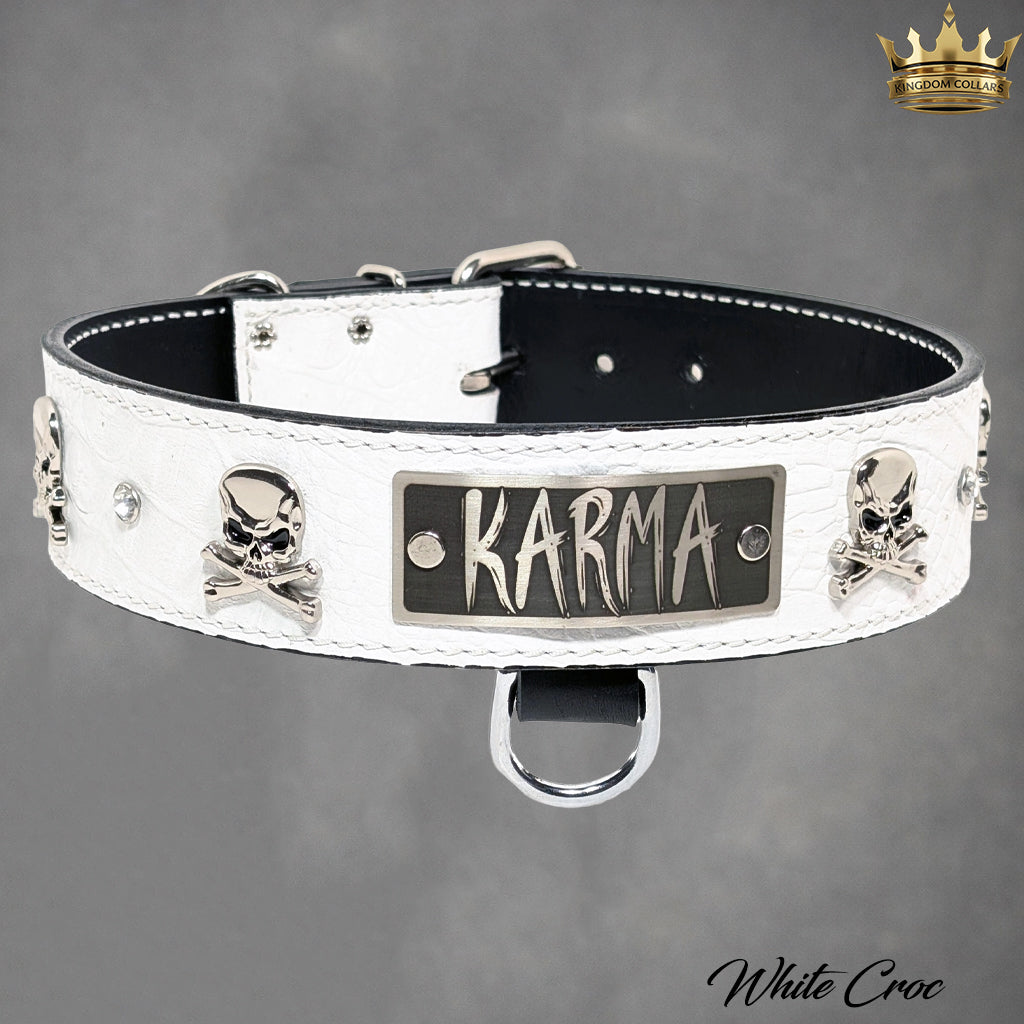 A white crocodile-embossed leather dog collar with a black interior, silver skull and crossbones studs, rhinestones, and a central metal plate engraved with 'KARMA'.