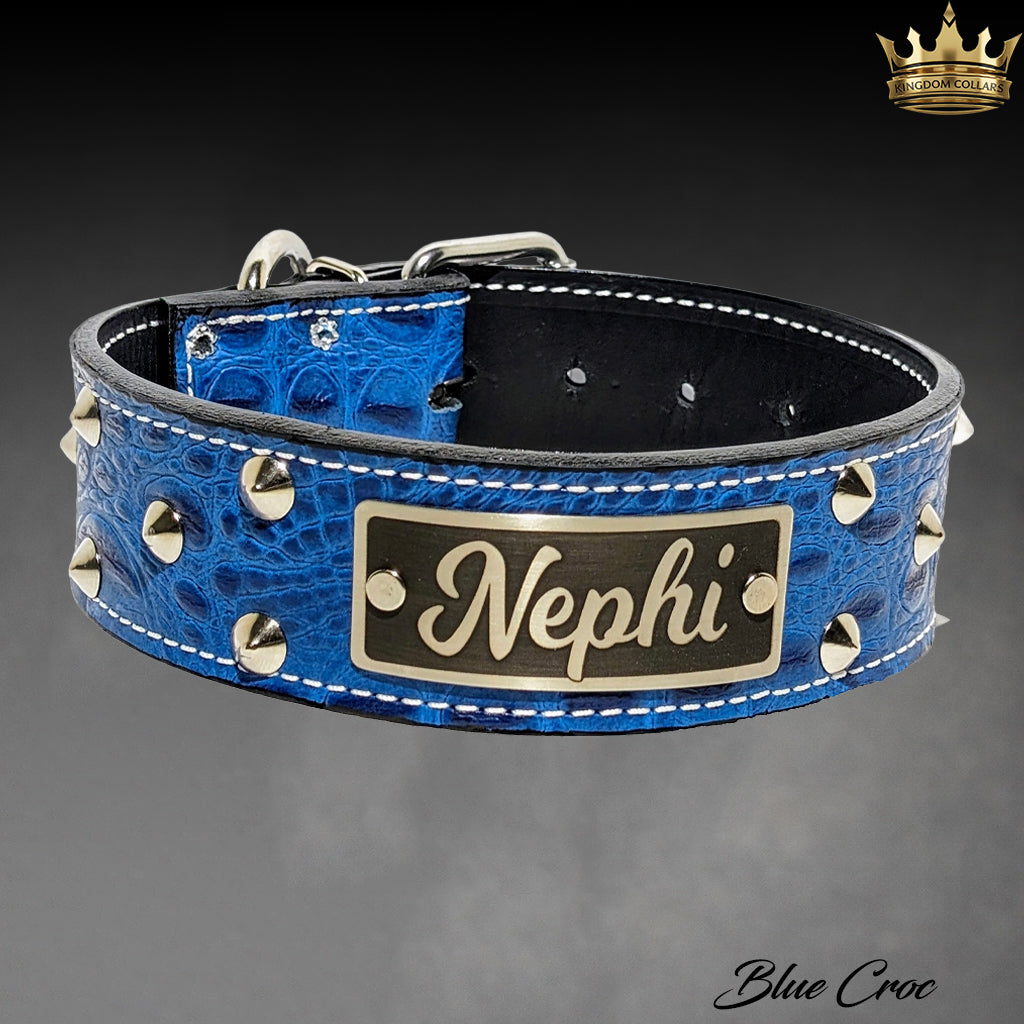 A blue crocodile-embossed leather dog collar with silver studs and a custom metal nameplate reading 'Nephi', shown on a dark gray gradient background.
