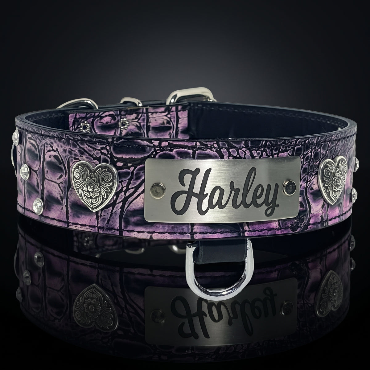 A purple and black crocodile-embossed leather pet collar with silver-tone metal hardware, including a central engraved metal plate that reads 'Harley', decorative heart medallions, and studs, shown on a dark reflective surface.