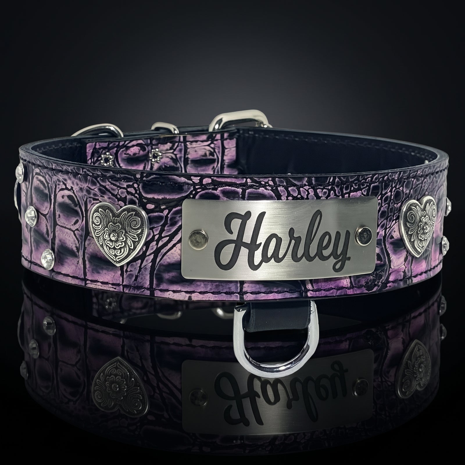 A purple and black crocodile-embossed leather pet collar with silver-tone metal hardware, including a central engraved metal plate that reads 'Harley', decorative heart medallions, and studs, shown on a dark reflective surface.