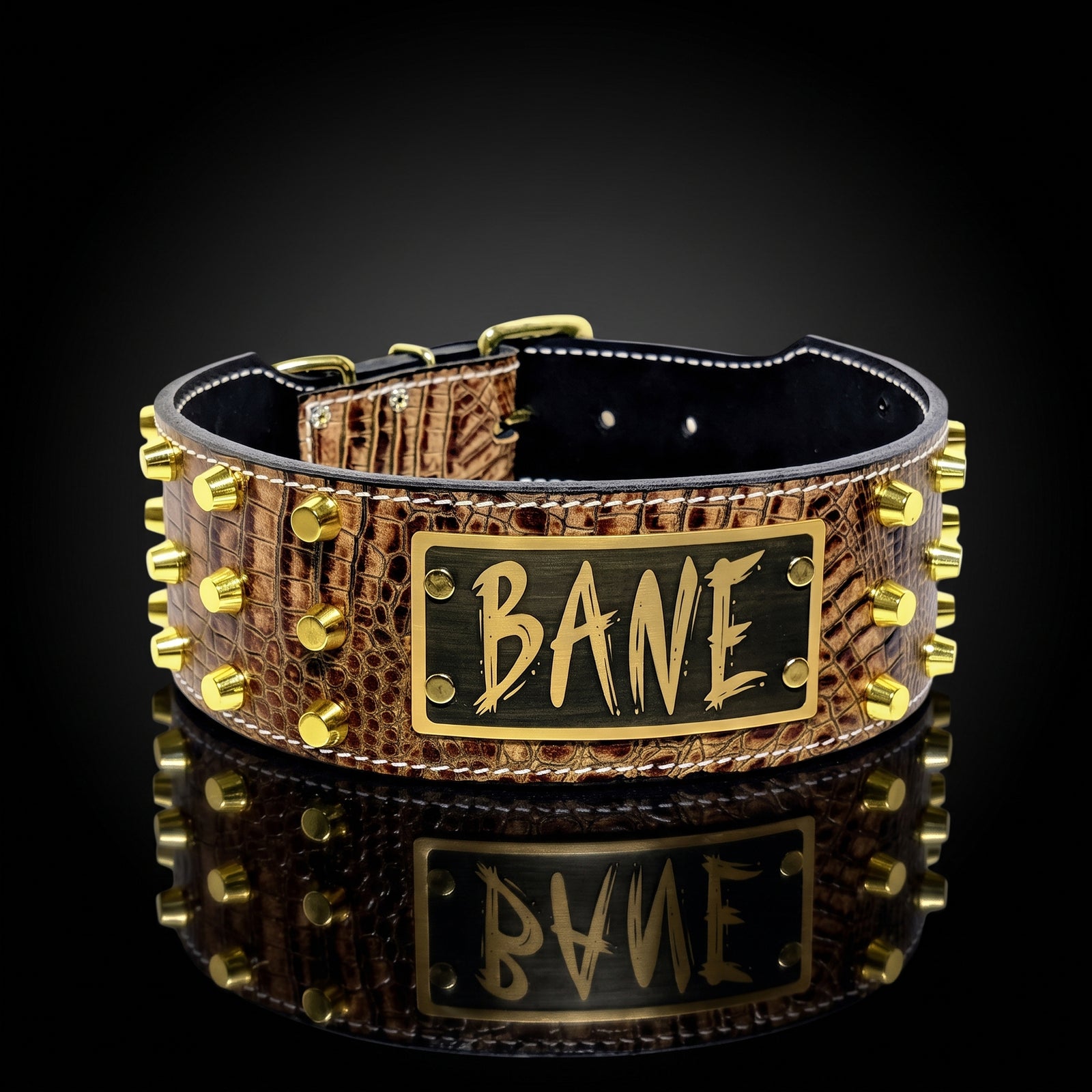 Cognac brown crocodile-embossed leather dog collar with three rows of gold cylinder studs and a brass nameplate engraved with "BANE" in gold graffiti-style lettering, displayed on a polished black stone surface with a soft reflection beneath, against a dark gradient background.