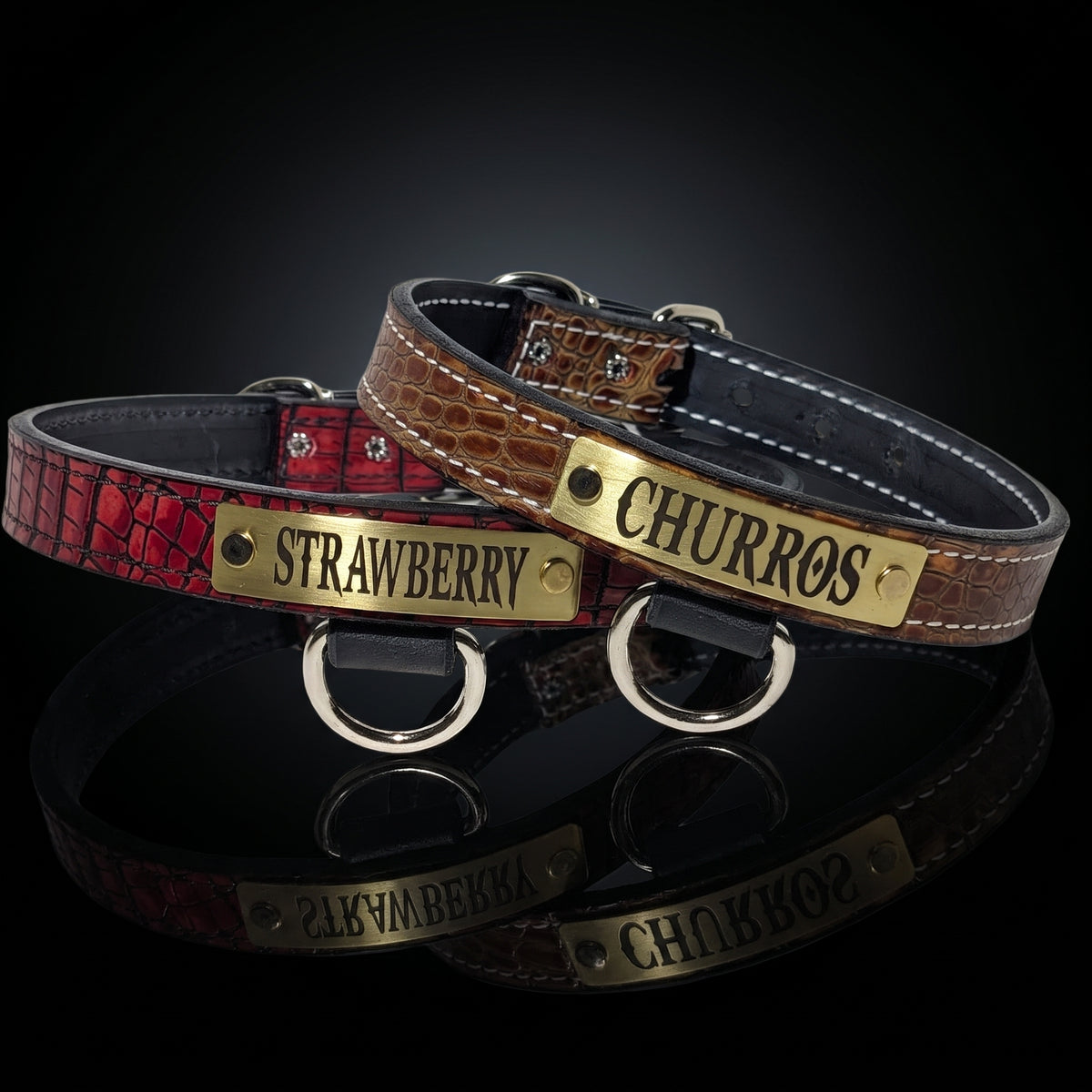 Two personalized dog collars displayed on a reflective black surface. One collar is textured red with a brass plate engraved 'STRAWBERRY', and the other is brown crocodile-embossed with a brass plate engraved 'CHURROS'.