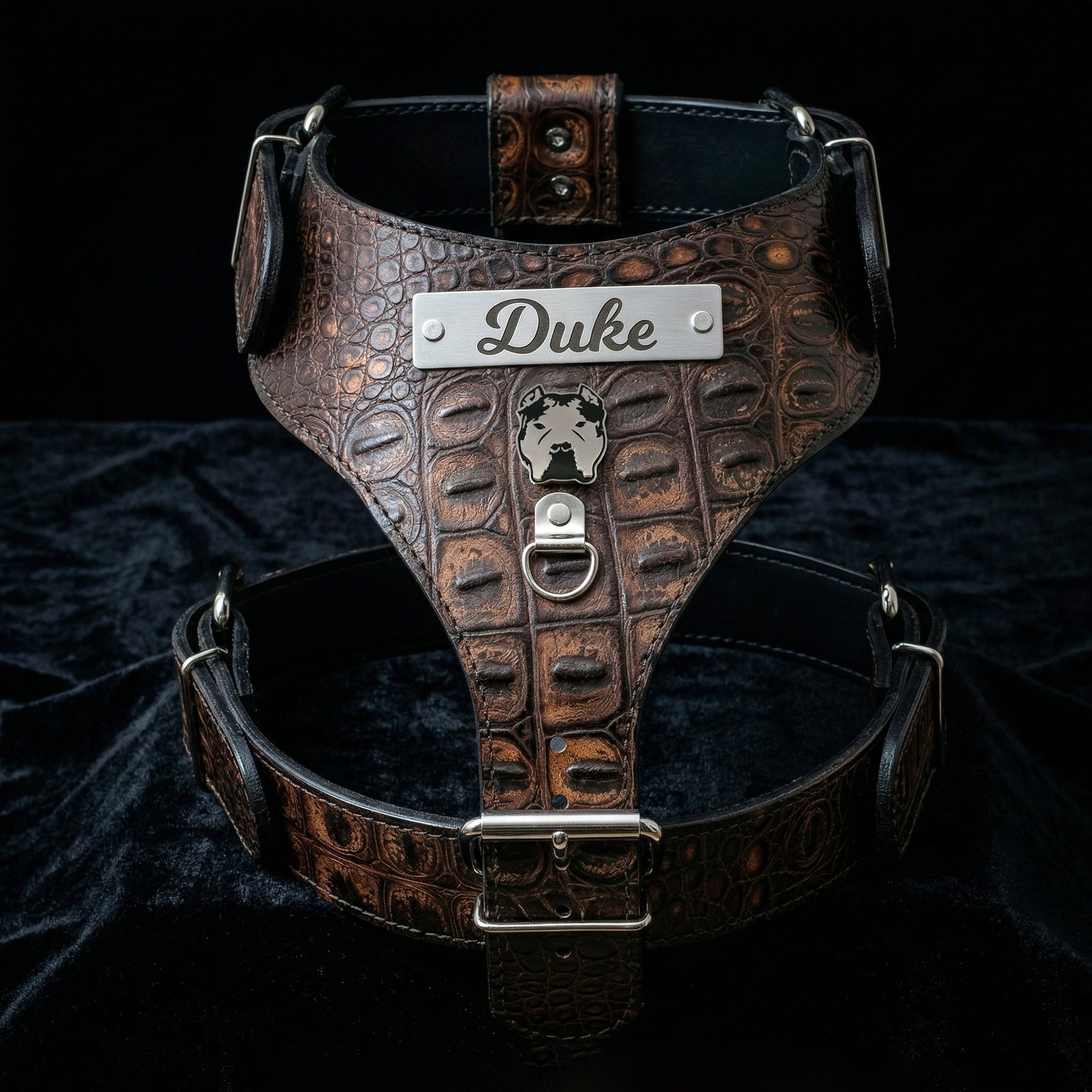 A brown embossed leather dog harness with a silver metal nameplate engraved 'Duke' and a small crest emblem, displayed on a black velvet fabric background.
