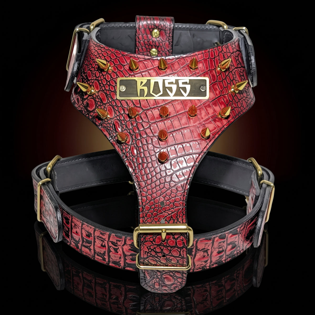 A red crocodile-embossed leather dog harness with gold-tone hardware, featuring a central brass plate engraved with 'BOSS' and gold spikes along the sides, set against a dark reflective background.