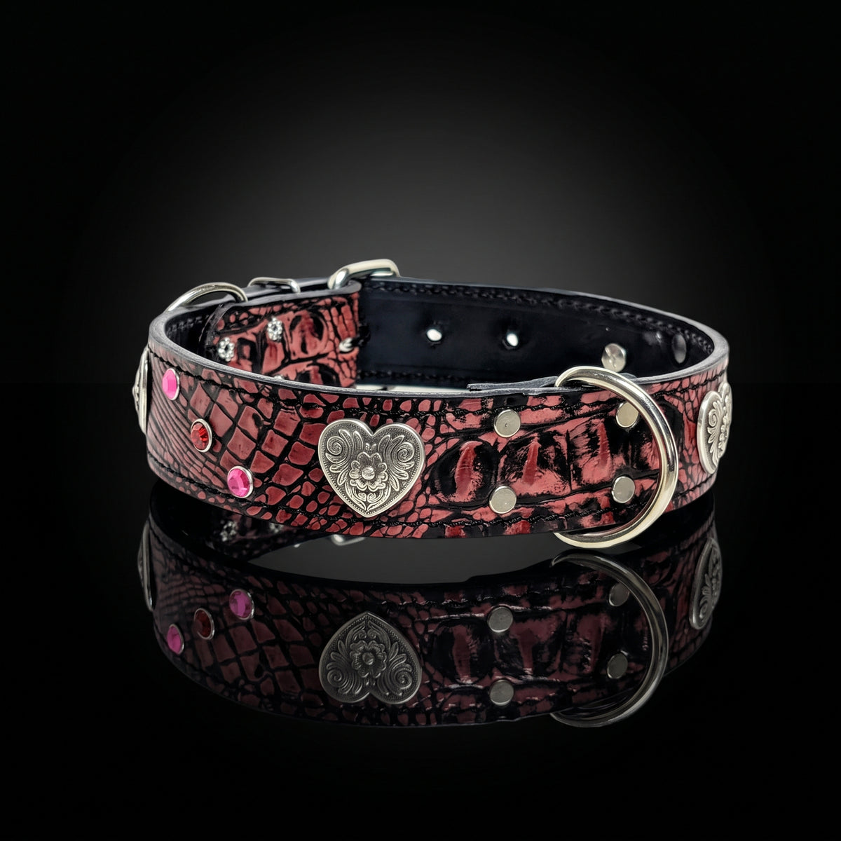 A gothic-style dog collar with a red and black crocodile-embossed pattern, featuring silver-tone metal studs, a central heart-shaped medallion with a skull design, and pink rhinestone accents, displayed on a reflective black background.