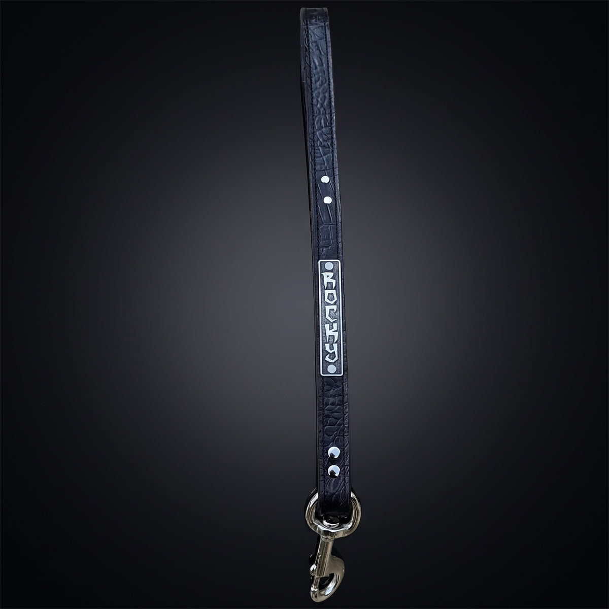 A black leather dog leash with silver hardware, featuring a silver metal nameplate engraved with the name 'JERSEY' and silver studs, shown against a dark gradient background.
