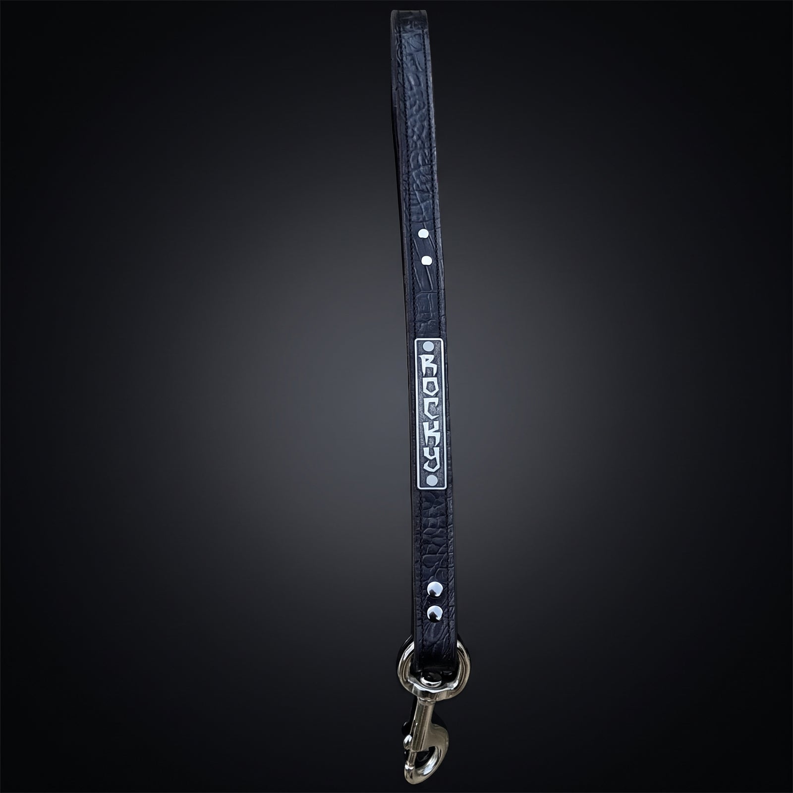 A black leather dog leash with silver hardware, featuring a silver metal nameplate engraved with the name 'JERSEY' and silver studs, shown against a dark gradient background.