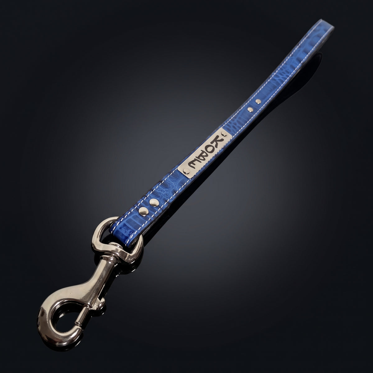 A blue crocodile-embossed leather strap with silver hardware, featuring a silver metal plate engraved with the brand name 'KOBE' and a silver swivel snap hook clasp, set against a dark gradient background.
