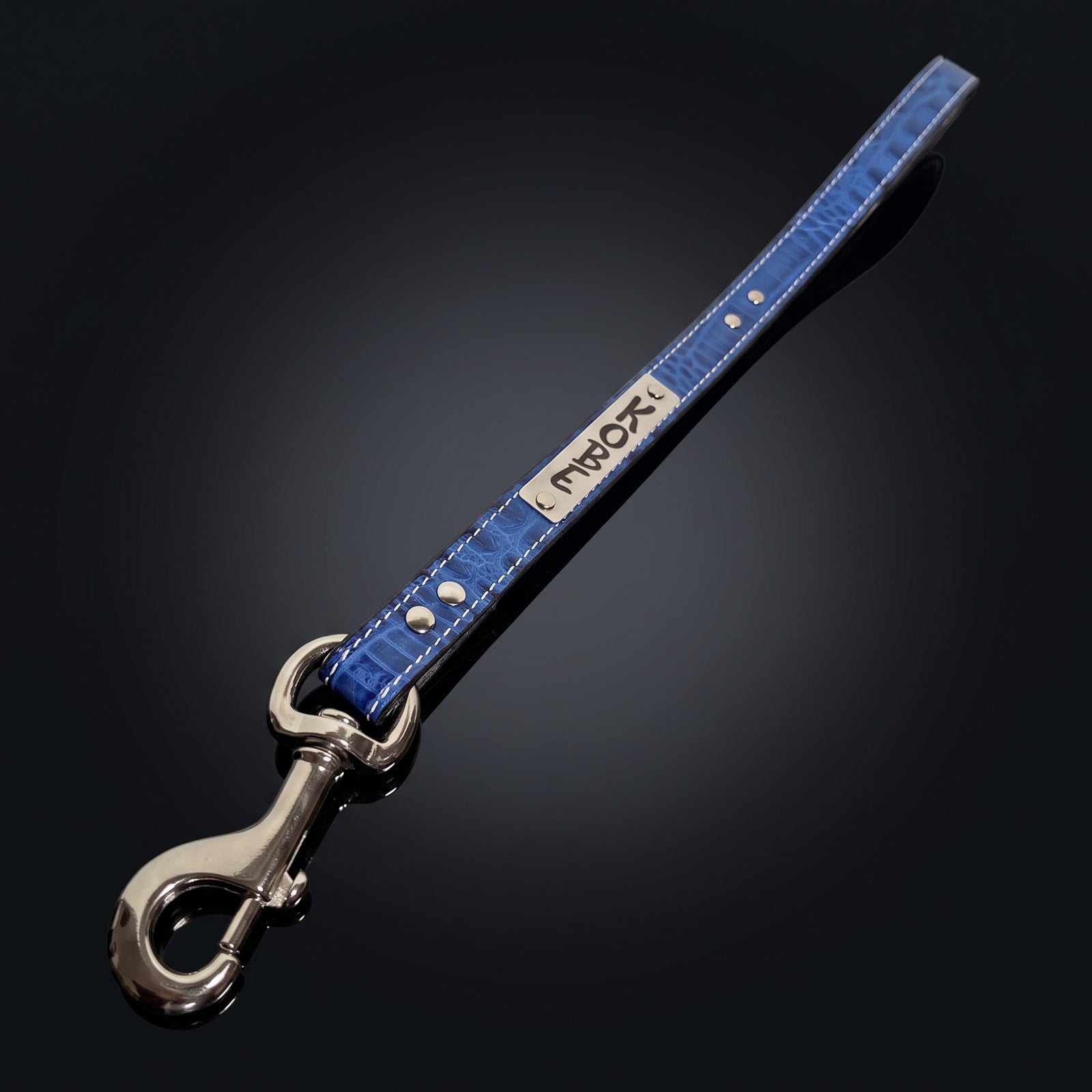 A blue crocodile-embossed leather strap with silver hardware, featuring a silver metal plate engraved with the brand name 'KOBE' and a silver swivel snap hook clasp, set against a dark gradient background.