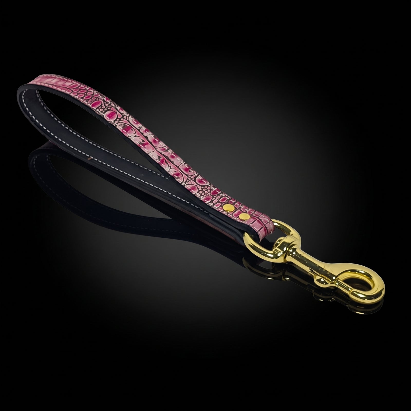 A pink patterned leather dog leash with a black underside, white stitching, and a gold-tone metal clasp, displayed on a reflective black background.
