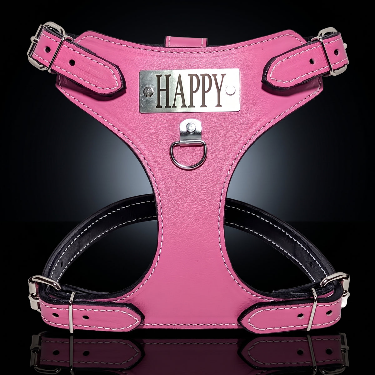 A pink leather dog harness with silver hardware, featuring a central metal plate engraved with the word 'HAPPY' and a black underbust band, set against a dark reflective background.