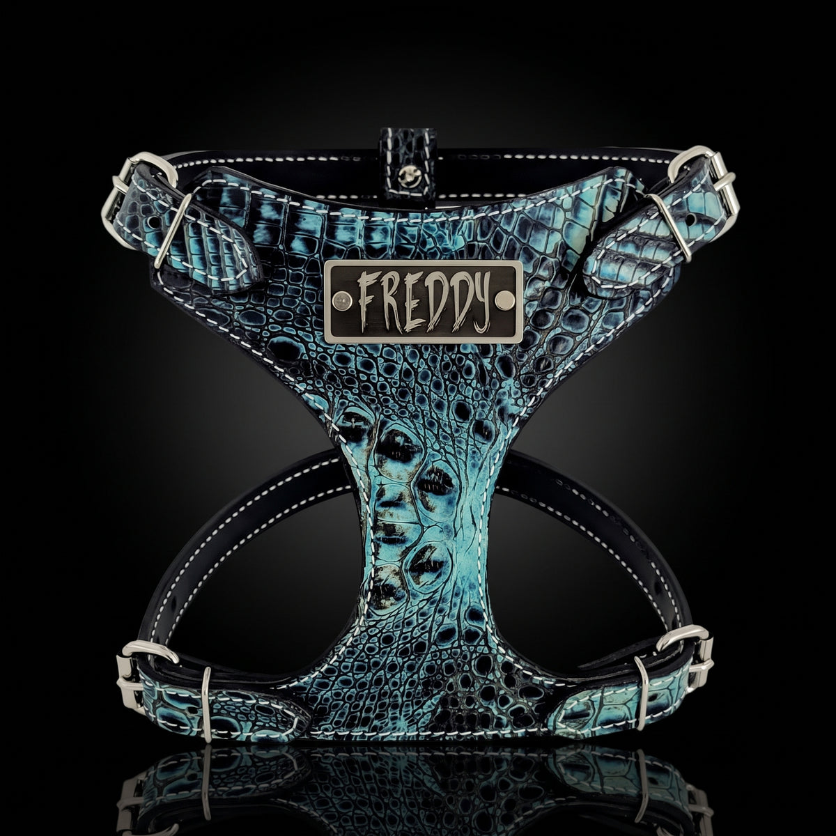 A dog harness with a textured turquoise and black crocodile pattern, featuring a central metal plate engraved with the name 'FREDDY'.