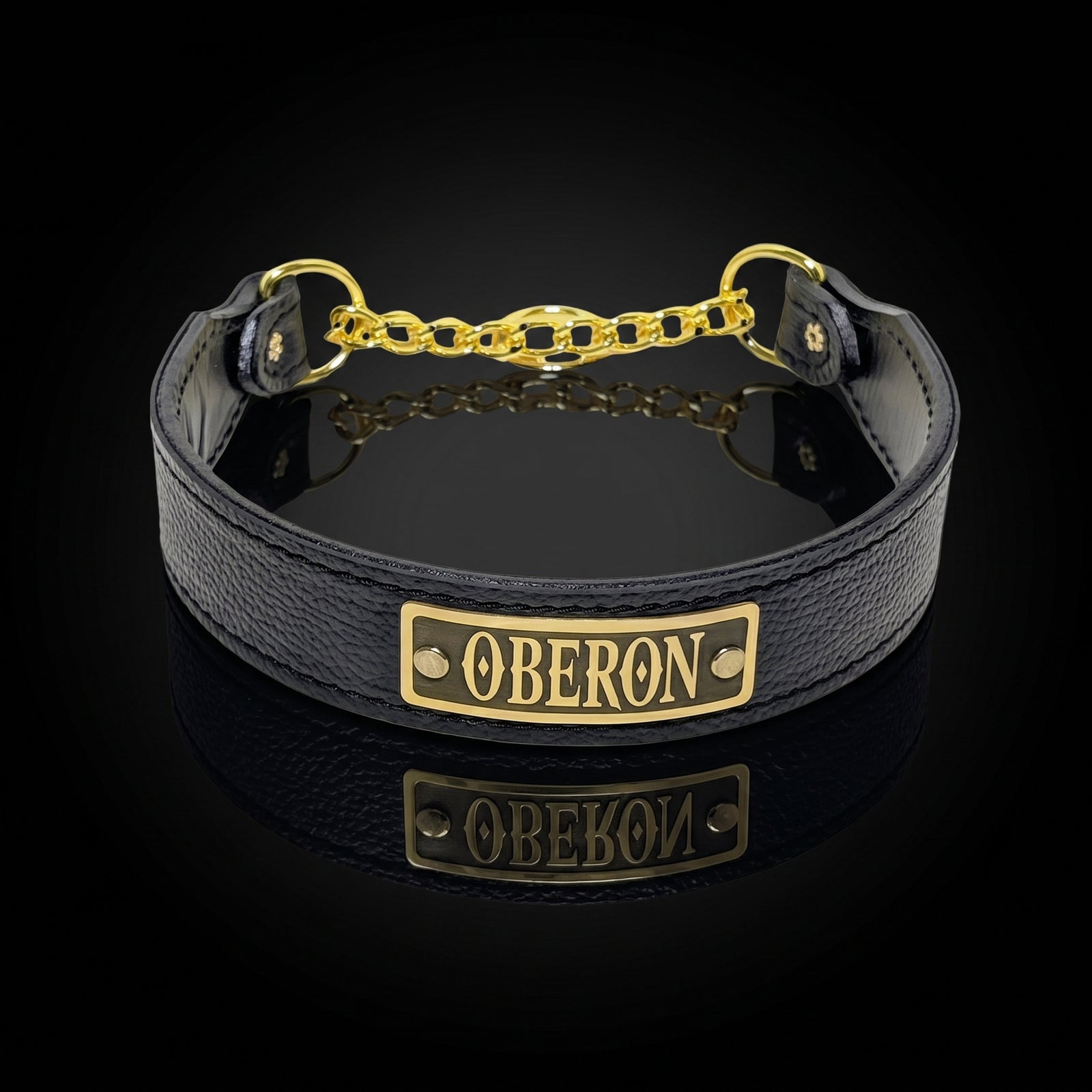 A black leather collar with a gold-tone metal plate engraved with 'OBERON' and a gold chain extender, displayed on a reflective black surface.