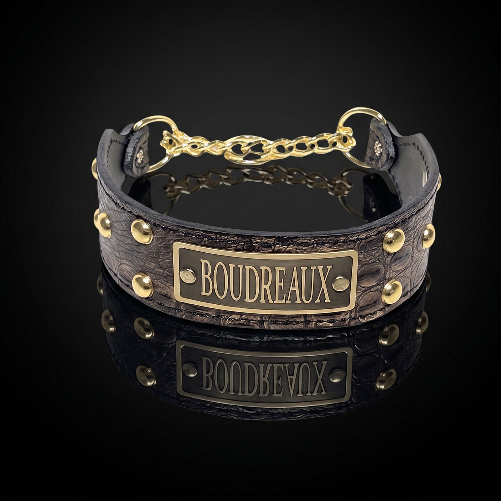A dark brown textured leather collar with gold-tone hardware, featuring a central brass plate engraved with the name 'BOUDREAUX' and a short chain connector, shown on a reflective black background.