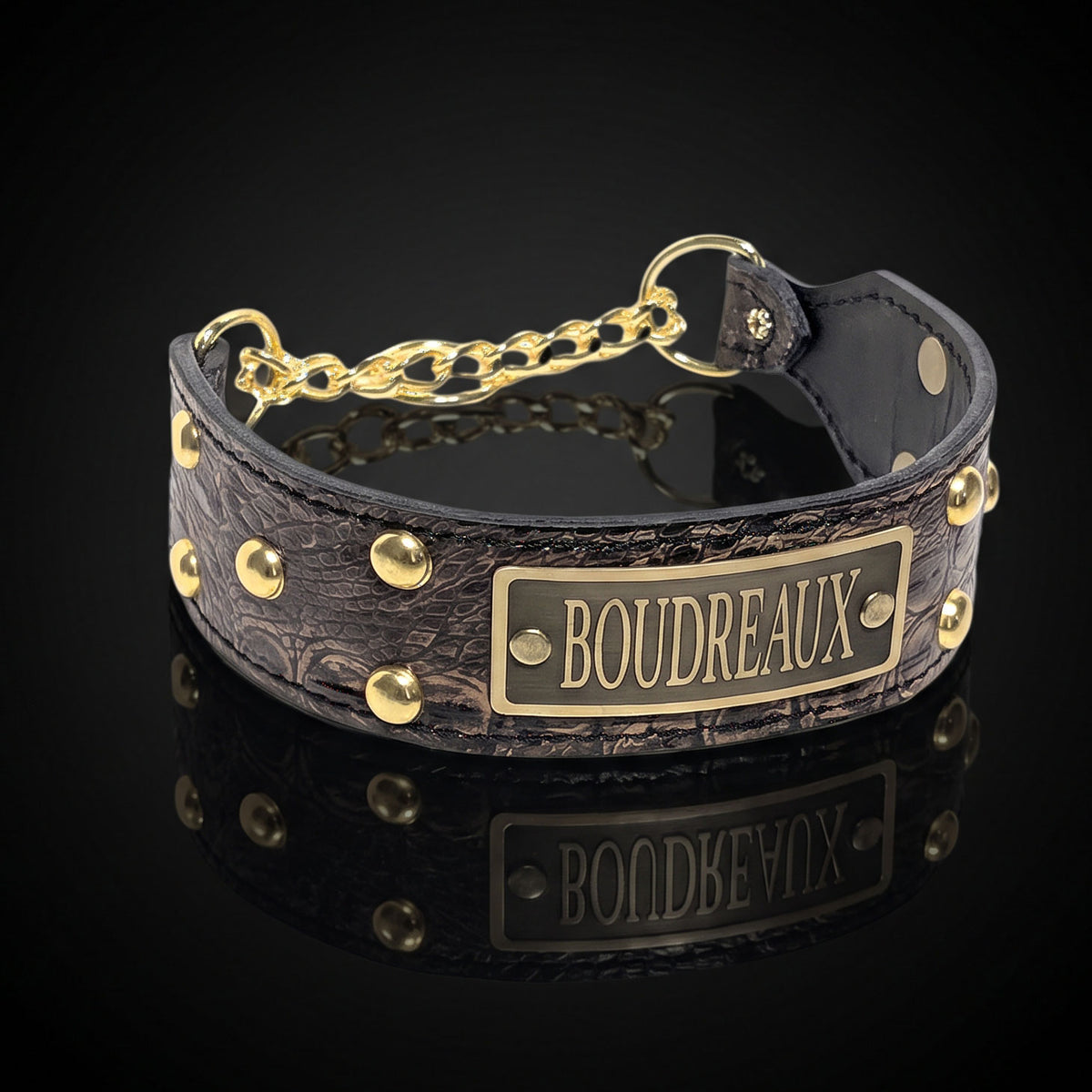 A dark brown textured leather collar with gold-tone hardware, featuring a rectangular brass plate engraved with the name 'BOUDREAUX' and a short chain connector, shown on a reflective black background.