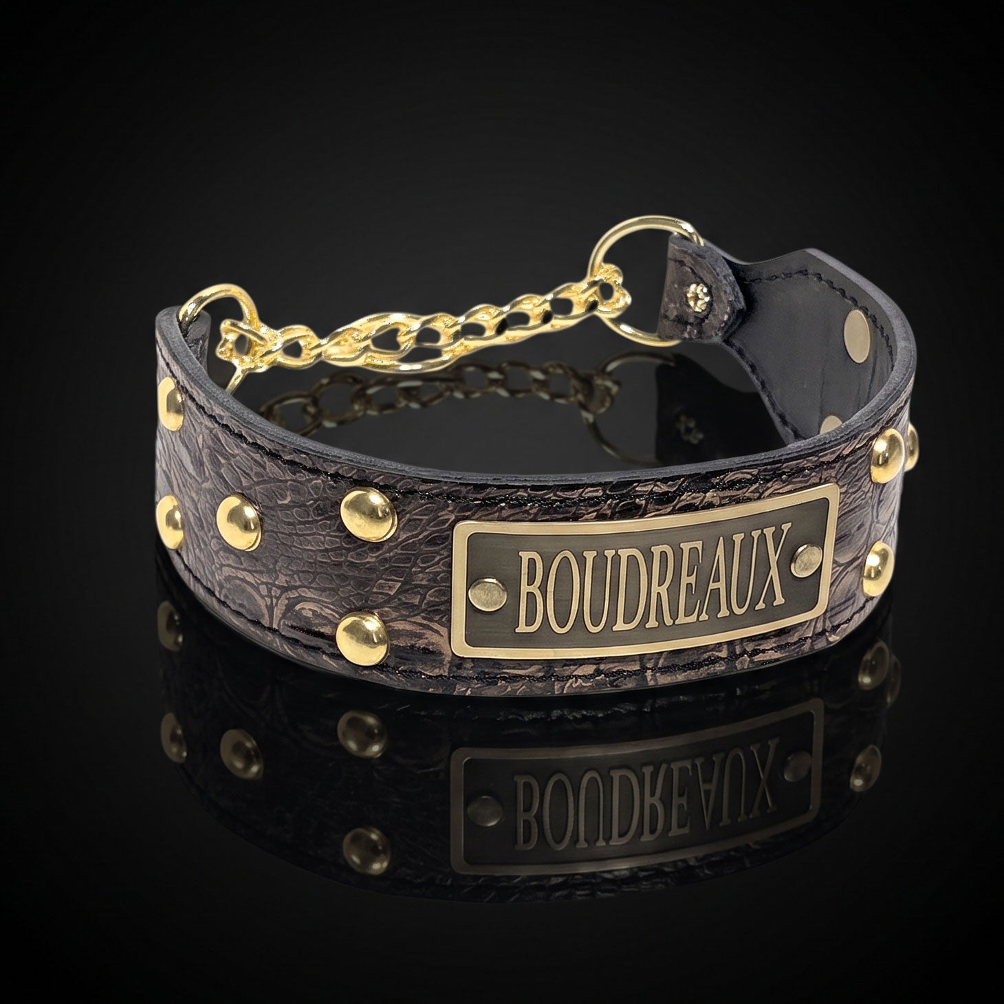 A dark brown textured leather collar with gold-tone hardware, featuring a rectangular brass plate engraved with the name 'BOUDREAUX' and a short chain connector, shown on a reflective black background.