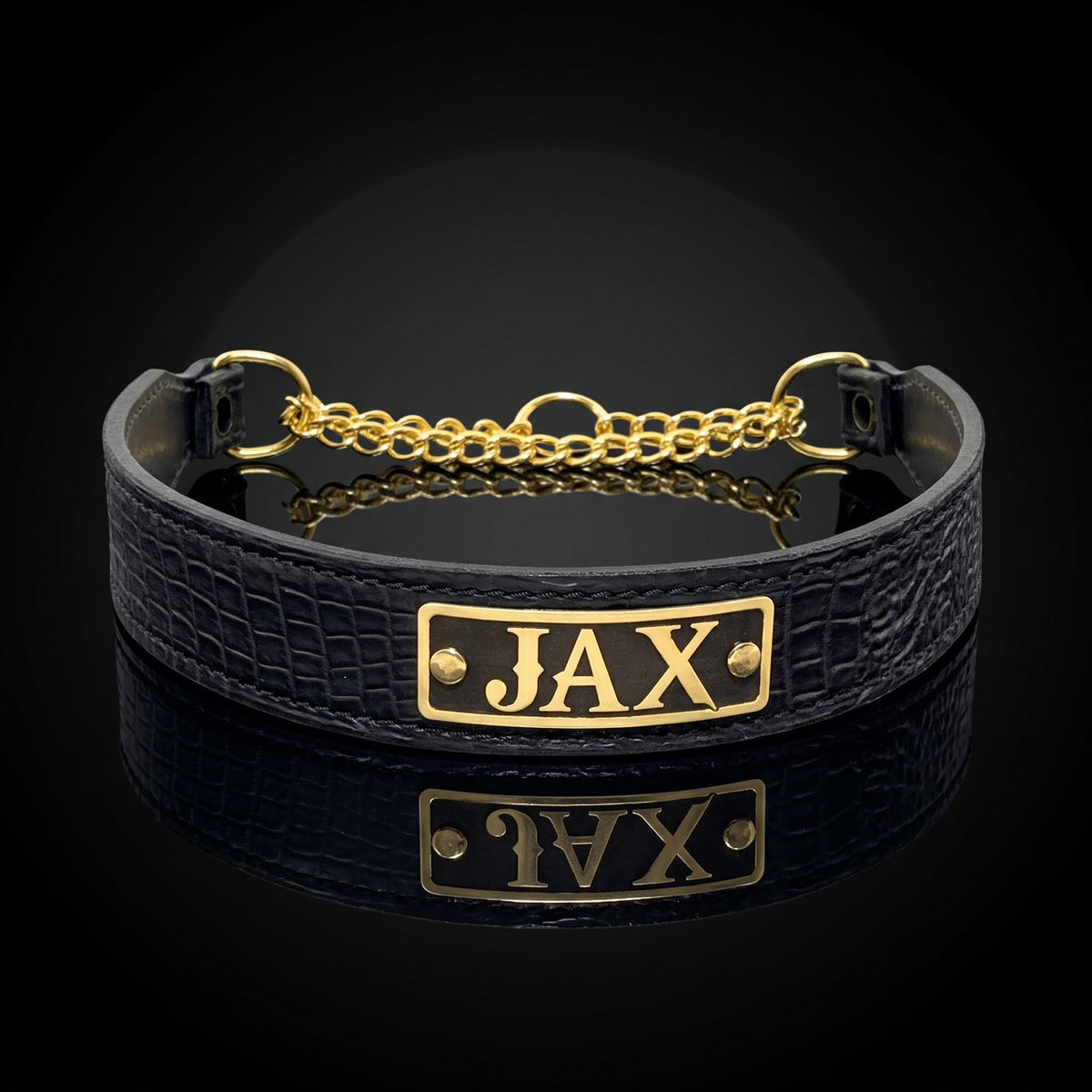 A black crocodile-embossed leather collar with a gold-plated metal plate engraved with the name 'JAX' and a gold chain closure, displayed on a reflective black surface.