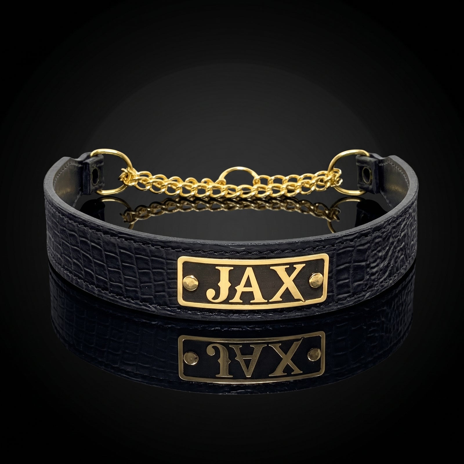 A black crocodile-embossed leather collar with a gold-plated metal plate engraved with the name 'JAX' and a gold chain closure, displayed on a reflective black surface.