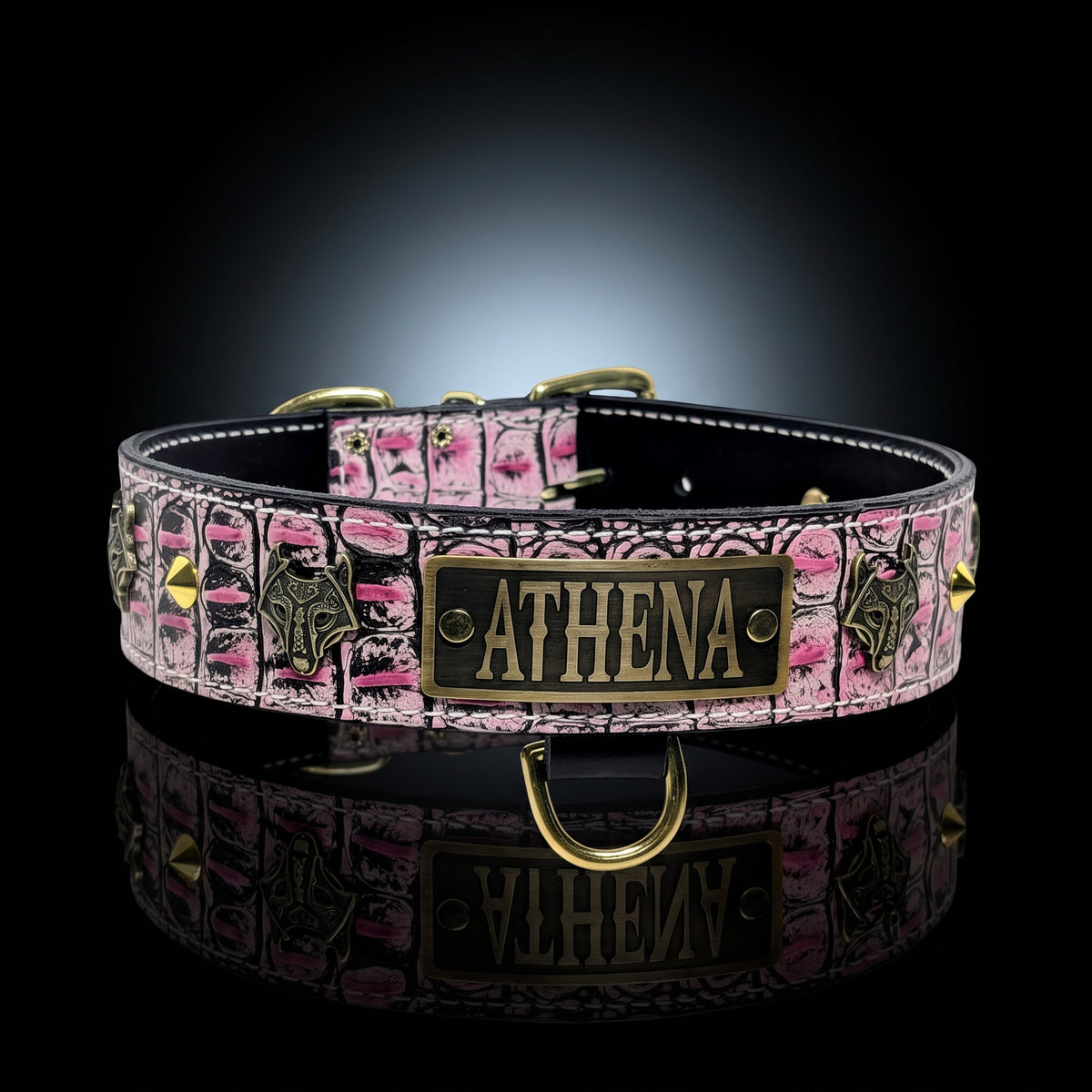 A custom pet collar with a pink and black patterned design, featuring a central brass plate engraved with the name 'ATHENA' and decorative bat motifs, displayed on a reflective black surface.