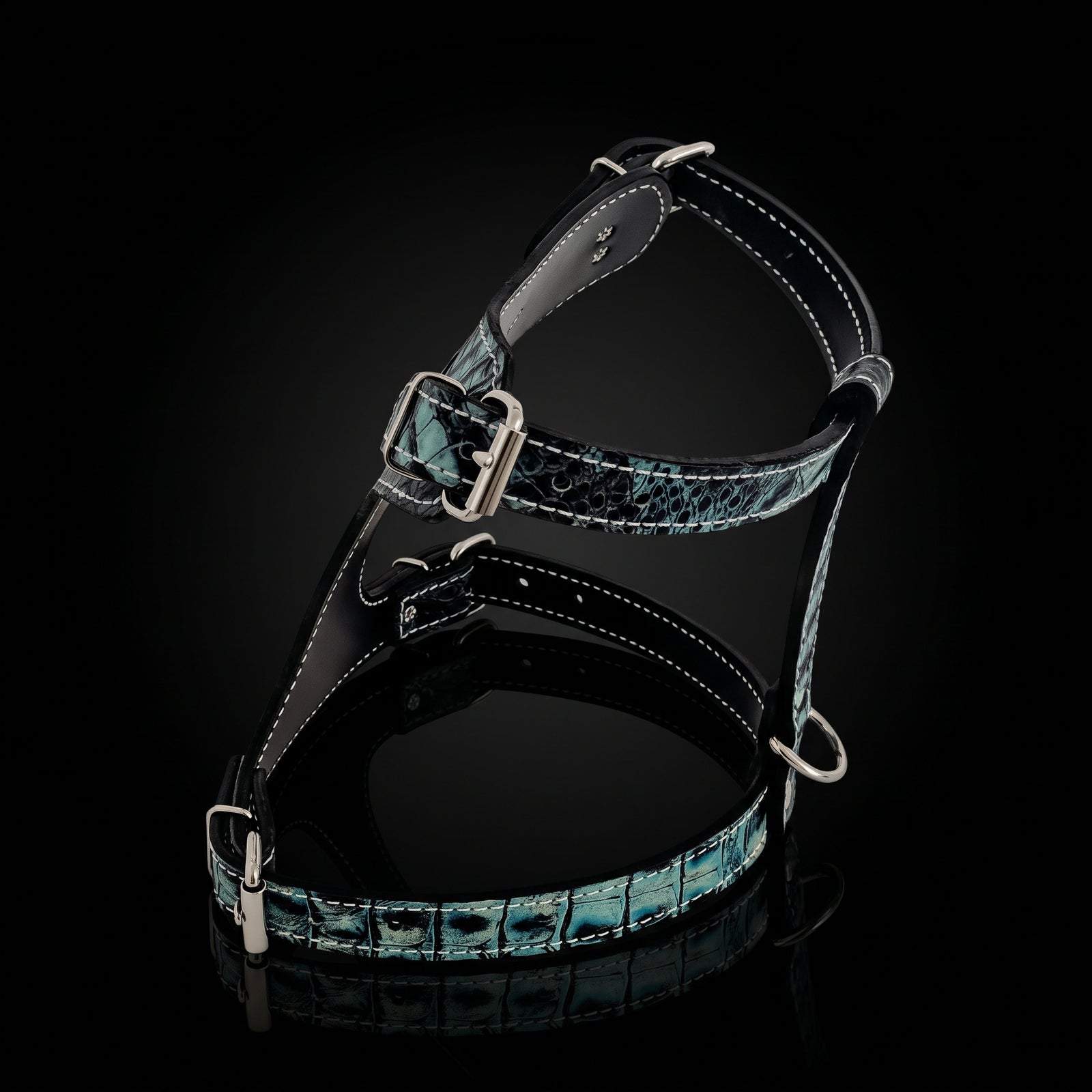 A black leather harness with a glossy blue-green crocodile or alligator print pattern, featuring silver-tone metal hardware and white contrast stitching, displayed on a reflective black background.
