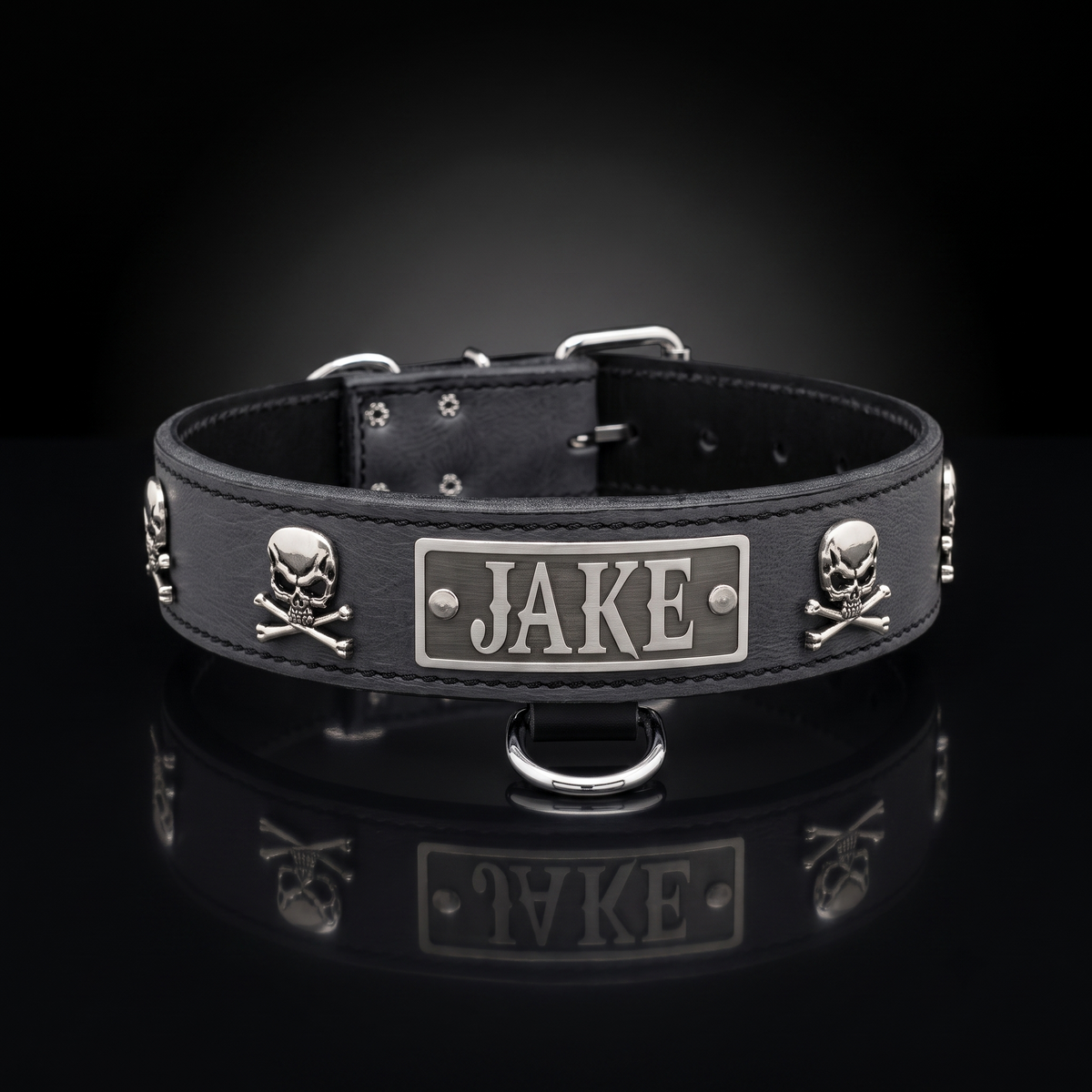 A black leather dog collar with silver metal hardware, featuring skull and crossbones studs and a central rectangular plate engraved with the name 'JAKE'. The collar is displayed on a reflective black surface against a dark gradient background.