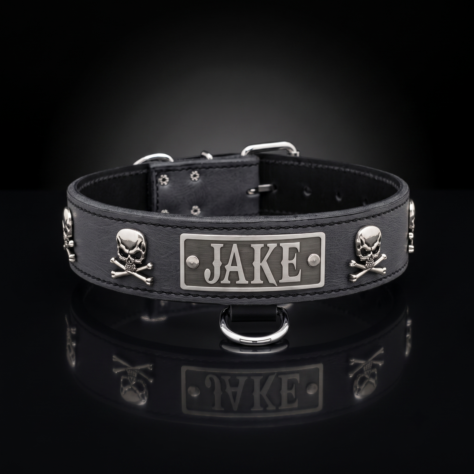 A black leather dog collar with silver metal hardware, featuring skull and crossbones studs and a central rectangular plate engraved with the name 'JAKE'. The collar is displayed on a reflective black surface against a dark gradient background.