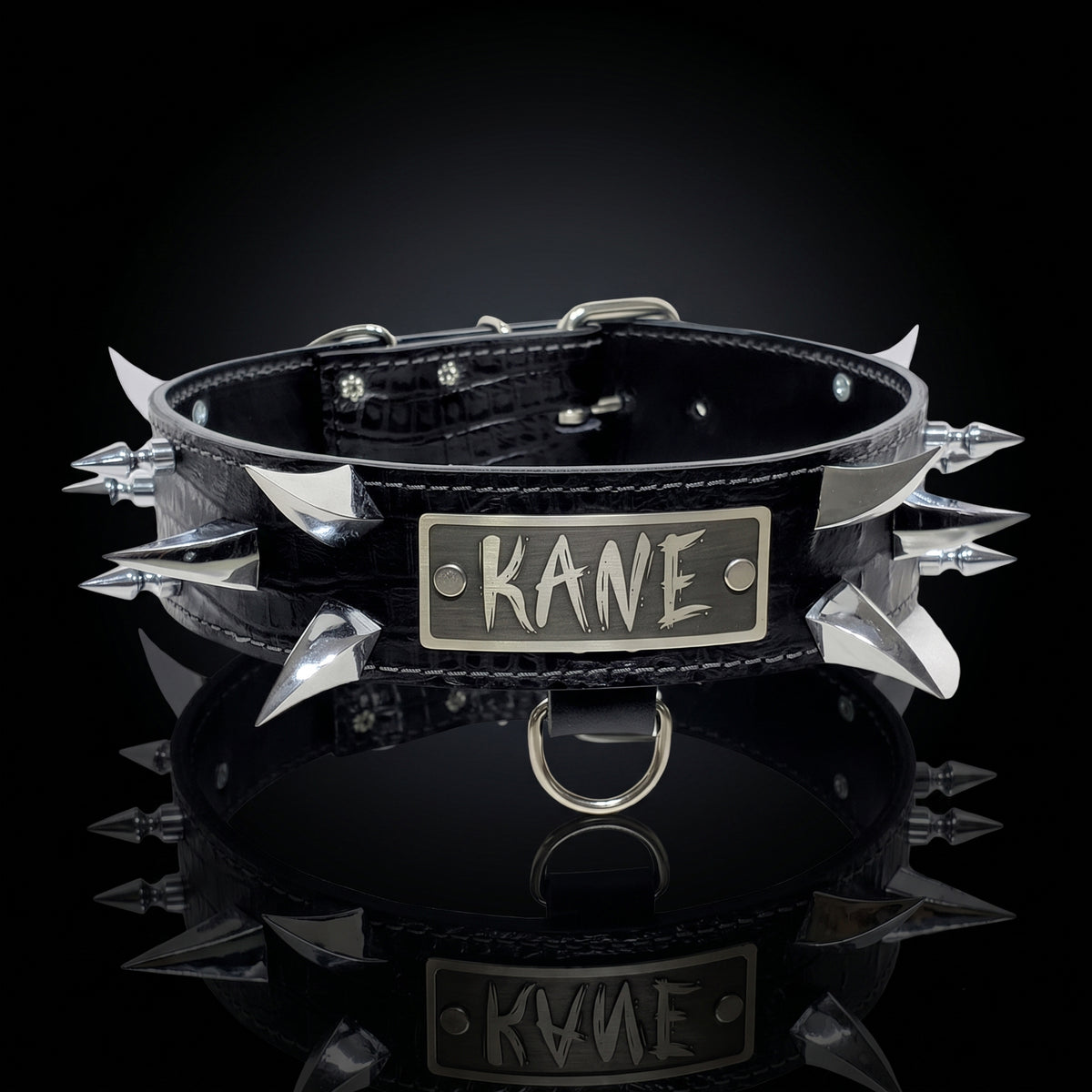 A black leather spiked collar with silver metal spikes and a rectangular silver plate engraved with the name 'KANE', shown on a reflective black surface against a dark background.