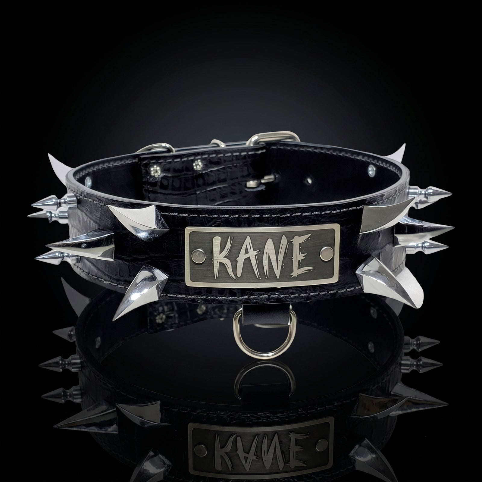 A black leather spiked collar with silver metal spikes and a rectangular silver plate engraved with the name 'KANE', shown on a reflective black surface against a dark background.