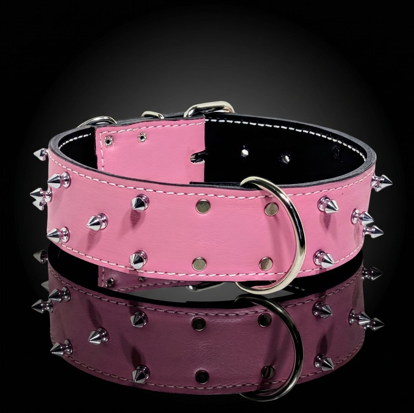 A pink leather dog collar with silver metal spikes and a large silver D-ring, displayed on a reflective black surface.