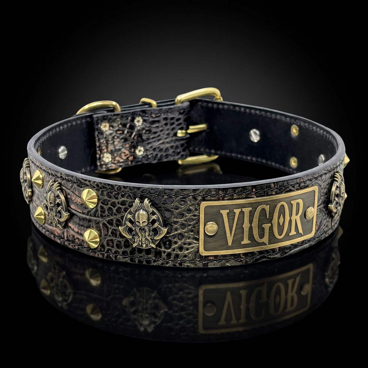 A black crocodile-embossed leather collar with antique gold-tone hardware, featuring a rectangular gold plate engraved with 'VIGOR' and decorative skull medallions, shown on a reflective black background.